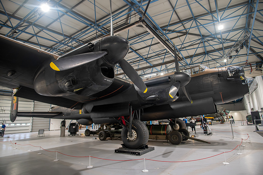 The image depicts a large, vintage military aircraft housed in a spacious hangar. The aircraft, painted in black, features distinctive yellow markings on its engines and nose. The hangar has a high ceiling with exposed blue metal beams and is well-lit with overhead lights. The aircraft is elevated on stands, and various maintenance equipment and tools are visible around it. The area is cordoned off with red ropes and stanchions, indicating it might be part of a museum or exhibition.