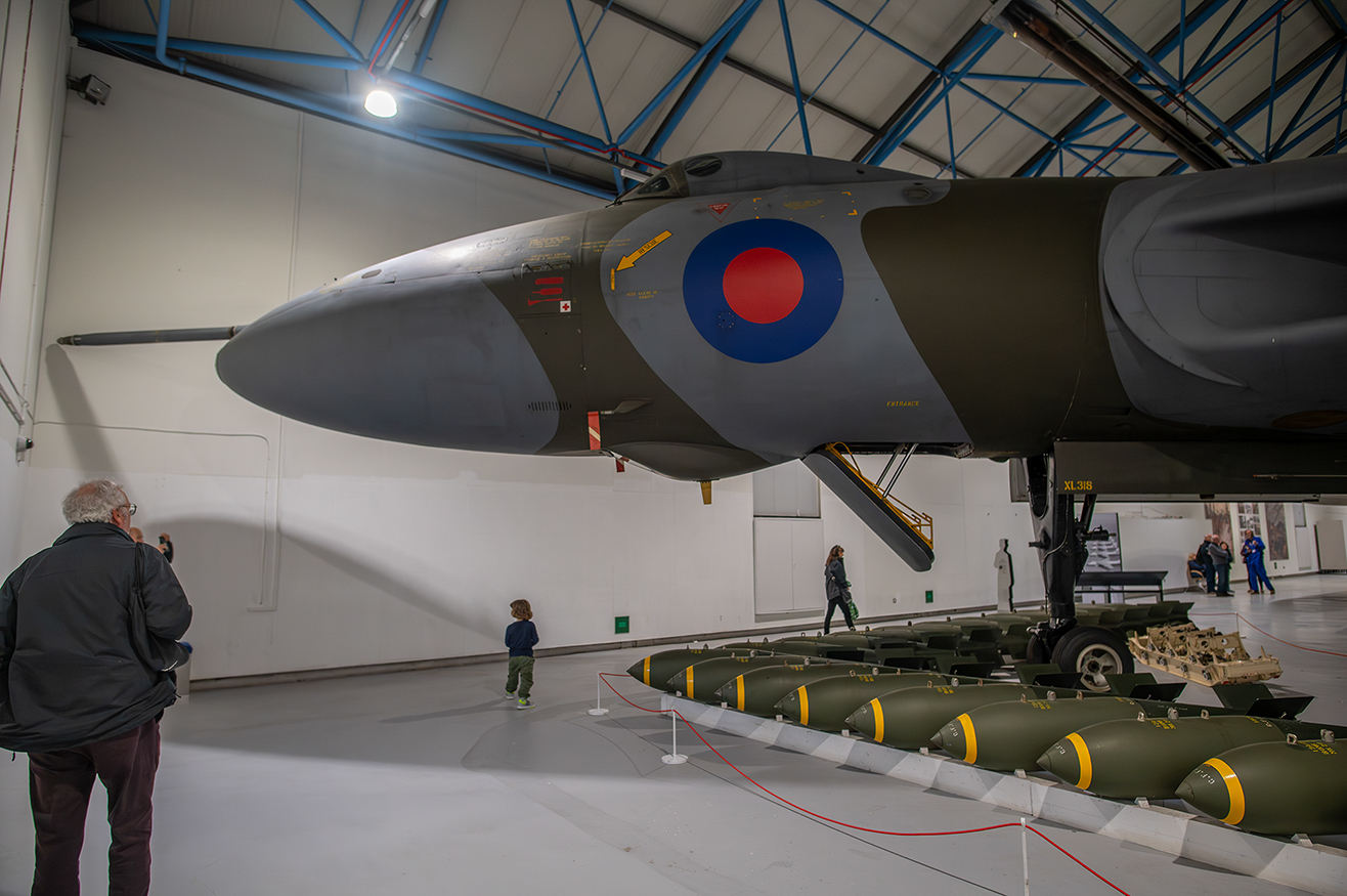 The image shows an aircraft on display in a museum setting, with a circular red, white, and blue roundel on its side. Several bombs are arranged on the ground in front of the aircraft. Visitors, including a child, are observing the exhibit. The aircraft is suspended, and its landing gear is down. The setting appears to be indoors with a high ceiling and industrial lighting.