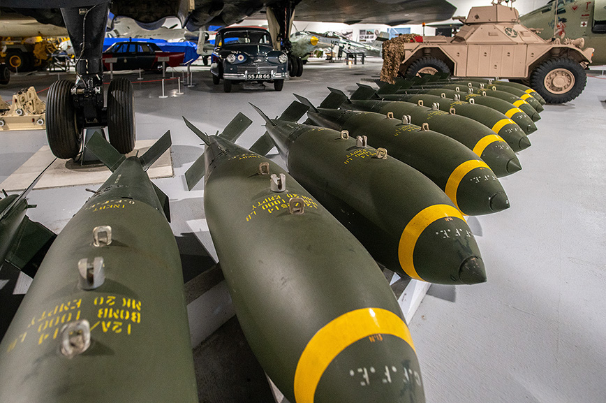 The image shows a collection of historical military bombs displayed in a museum setting. The bombs are lined up in a row, with various markings and labels on them. In the background, there are other military vehicles and artifacts, including an aircraft and a vintage car. The display appears to be part of a larger exhibition showcasing military history.