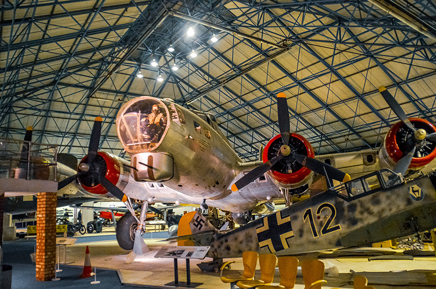 The image depicts a vintage military aircraft displayed in a museum setting. The aircraft, marked with the number 12 and a cross insignia, is a twin-engine, multi-propeller plane. It is elevated on supports, allowing viewers to see underneath it. The interior of the cockpit is visible, and the plane is surrounded by informational displays and barriers, indicating it is part of an exhibit. The setting appears to be a large, well-lit hangar with other exhibits visible in the background.