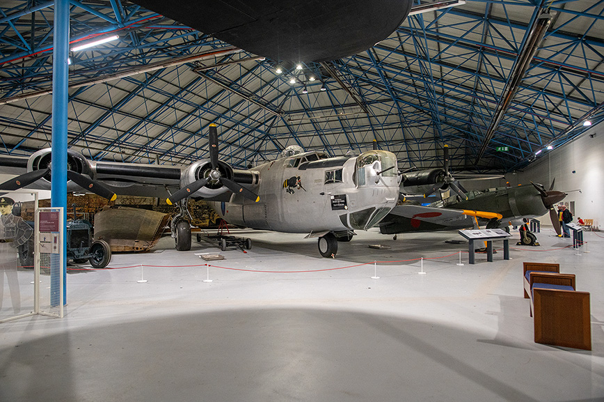 The document describes an image of an aircraft hangar housing several vintage military aircraft. The hangar has a large, open space with a high ceiling supported by a blue truss structure. The central focus is a large, gray, four-engine bomber with distinctive markings and a turret on top. Other aircraft, including a smaller plane with a red and white nose, are also visible. The area is cordoned off with red ropes and stanchions, and there are informational displays and a motorcycle on the left side of the image. The overall atmosphere suggests a museum or exhibition setting dedicated to aviation history.