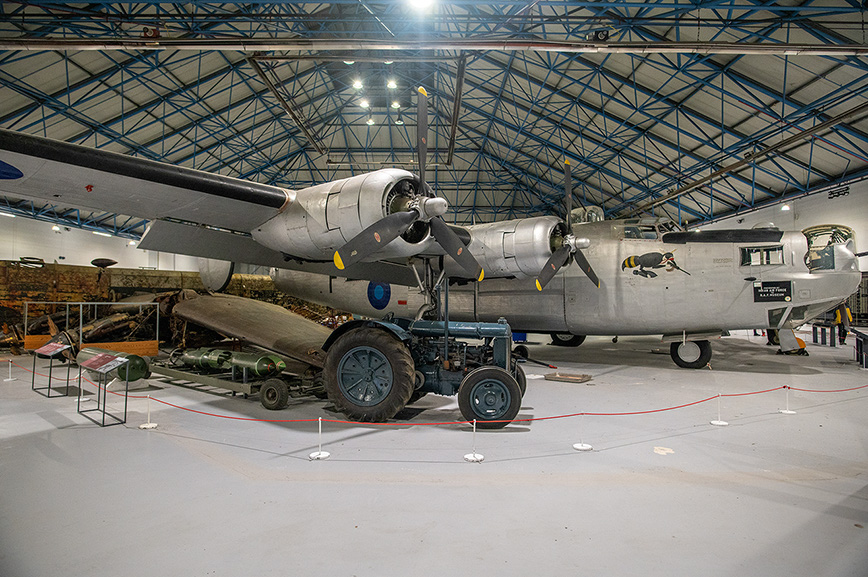 The image depicts a vintage military aircraft displayed in a museum setting. The aircraft, a twin-engine bomber, is shown with its bomb bay doors open, revealing several bombs. The plane is housed in a large hangar with a high, arched ceiling supported by metal trusses. The floor is clean and marked with red ropes to guide visitors and protect the exhibits. Additional historical artifacts and informational displays are visible in the background.