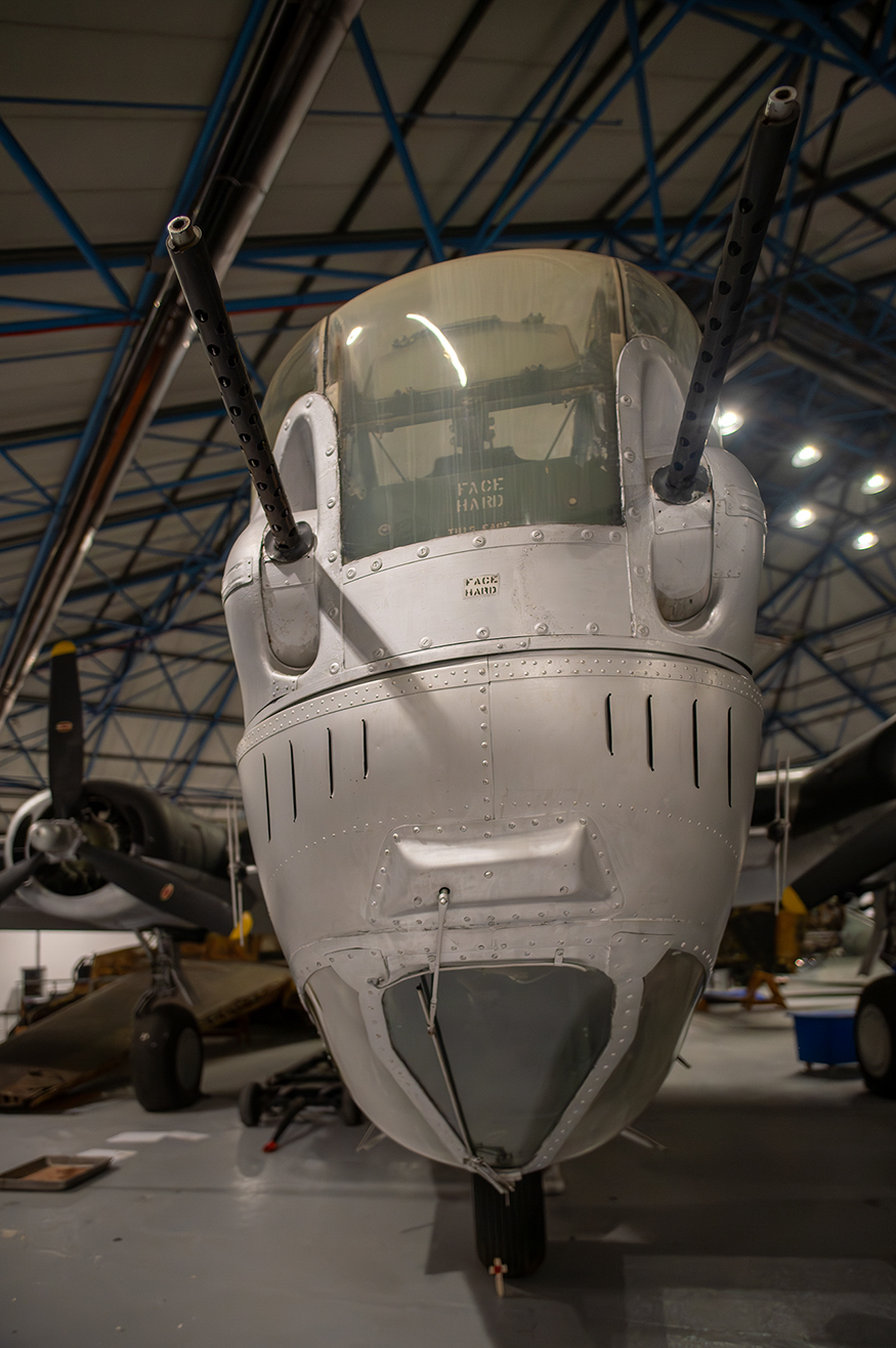 The image shows the front view of a vintage military aircraft, specifically a B-24 Liberator bomber. The aircraft's nose is prominently displayed, featuring a clear bombarder's window with the words 'FACE HARD' written on it. The aircraft is housed in a large hangar with a high ceiling and industrial lighting. Other aircraft and equipment are visible in the background, indicating that this is likely a museum or an aviation display area.