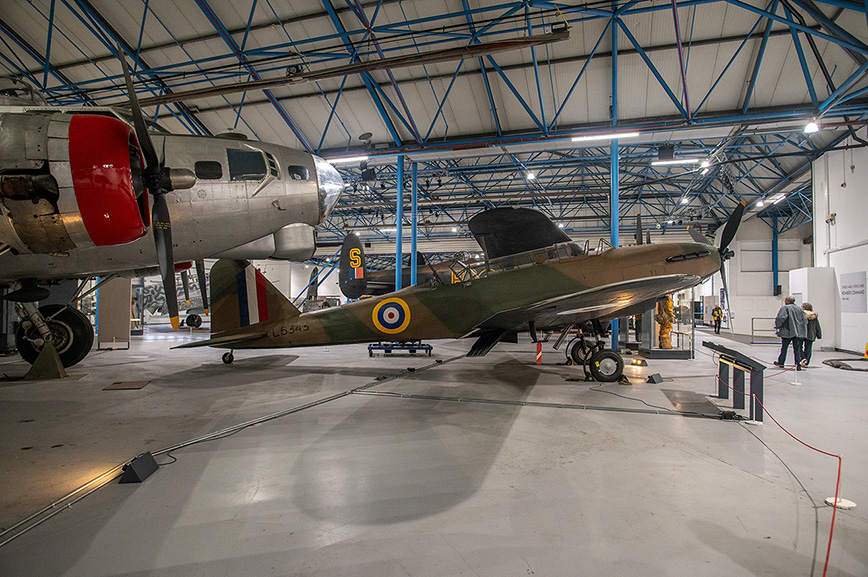 The image shows the interior of an aviation museum or hangar. Two vintage aircraft are prominently displayed: a silver and red multi-engine plane on the left and a green single-engine plane with a roundel marking on the right. The hangar has a high ceiling with blue structural elements and is well-lit. There are a few people visible in the background, one of whom appears to be a museum staff member in a yellow jacket. The overall scene suggests a well-maintained and organized exhibit space dedicated to preserving and showcasing historical aircraft.
