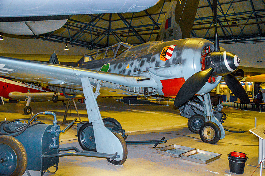 The image shows a vintage military aircraft displayed in a museum setting. The aircraft has a distinctive camouflage paint scheme with red, white, and green markings. It features a propeller at the front and is equipped with landing gear. The aircraft is positioned indoors, with other aircraft visible in the background. Various maintenance tools and equipment are scattered around the floor near the aircraft.
