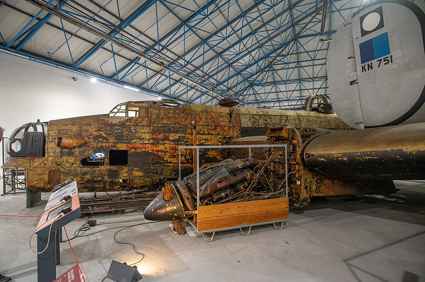 The image shows a vintage, rusted aircraft displayed in a museum-like setting. The aircraft is partially disassembled, with its engine and other components visible. The setting appears to be an indoor hangar with a high, blue-trussed ceiling. There is an information stand in front of the aircraft, likely providing details about its history and specifications. The aircraft's tail section shows the identification number 'KN 751'.