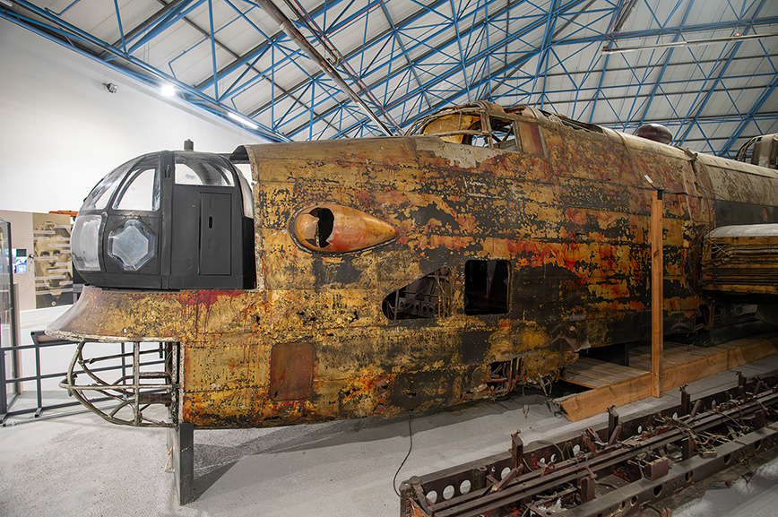 The image depicts an old, rusted locomotive inside a large, industrial building with a high, blue ceiling. The locomotive is heavily weathered, with visible rust and paint peeling off, and is supported by wooden beams. The front of the locomotive features a distinctive, rounded design with a large, clear windshield and a smaller, hexagonal window on the side. The setting suggests a museum or a restoration facility.