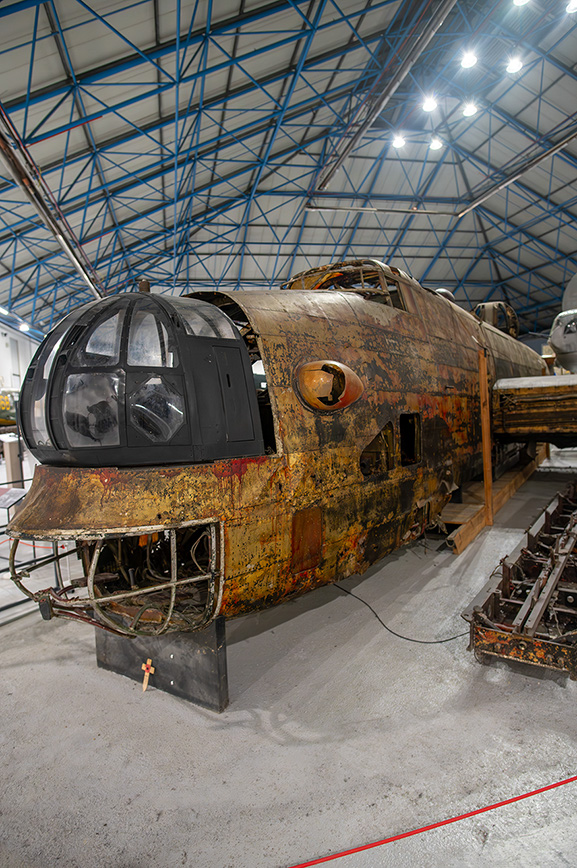 The image shows a rusted and aged aircraft, likely a historical or vintage model, displayed in a large hangar with a blue metal framework roof. The aircraft's exterior is heavily corroded, indicating it has not been used or maintained for a long time. The cockpit is prominently visible with a transparent dome, and the overall structure appears to be in a state of disrepair. The setting suggests it might be part of a museum or a storage facility for old aircraft.