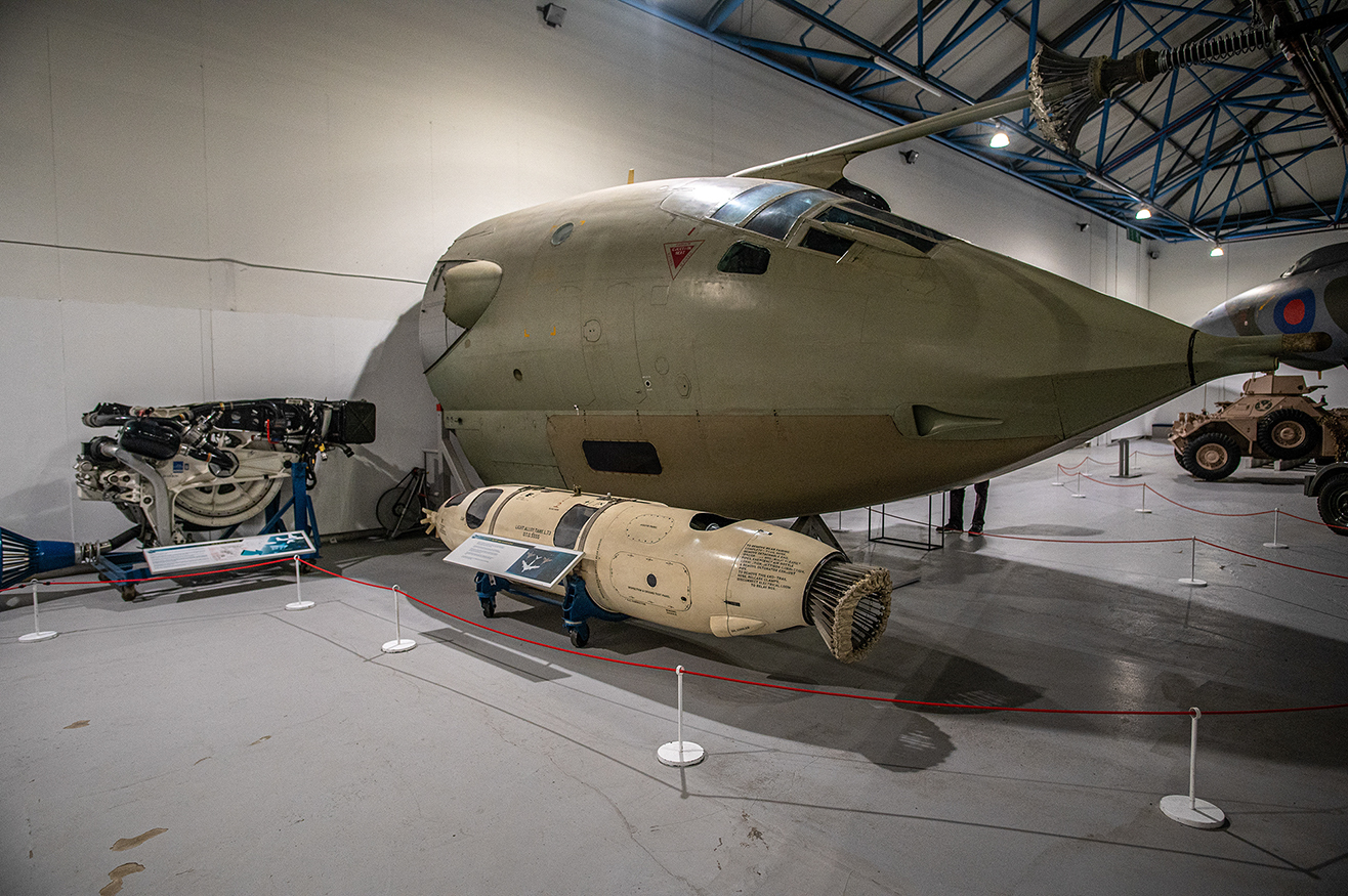 The image depicts a museum exhibit featuring a large military aircraft, likely a bomber, with a distinctive rounded nose. In front of the aircraft, there is a large bomb or missile on display. Additionally, an aircraft engine is exhibited nearby. The exhibit is cordoned off with red ropes and stanchions, and there are informational placards in front of each item, providing details about the artifacts.