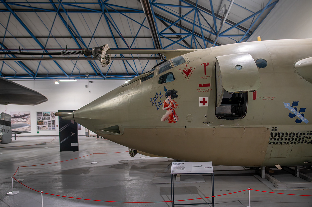 The image depicts the nose section of a military aircraft displayed in a museum. The aircraft is adorned with various markings, including a pin-up girl illustration labeled 'Lucky Lou', a red cross, and several warning labels. The aircraft is housed in a spacious, well-lit hangar with a high, arched ceiling supported by blue framework. There are informational displays and other aircraft parts visible in the background. The aircraft is cordoned off with red ropes and white stands, indicating it is part of an exhibit.