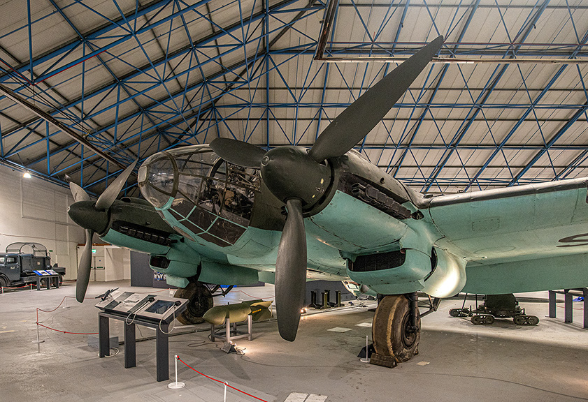 The image shows a vintage military aircraft displayed in a museum setting. The aircraft is a twin-engine, propeller-driven bomber with a distinctive green paint scheme. It is housed in a large hangar with a high, arched roof supported by blue trusses. Informational displays and a bomb are positioned nearby, providing context and details about the aircraft. The overall scene suggests a well-preserved historical exhibit.
