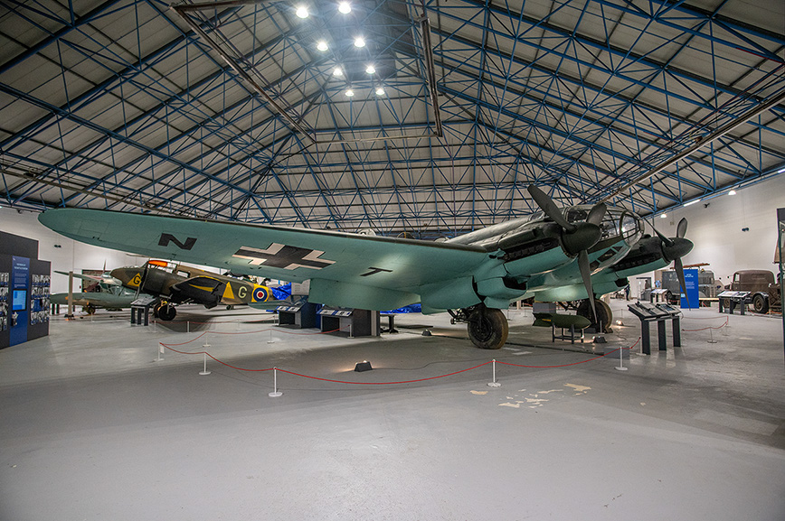 The image depicts an aircraft museum exhibit featuring a large, vintage military airplane prominently displayed in the center. The aircraft has a distinctive green color scheme with black markings. The museum's interior is spacious with a high, arched ceiling supported by a metal framework. Other aircraft and informational displays are visible in the background, indicating a collection of historical aviation artifacts.