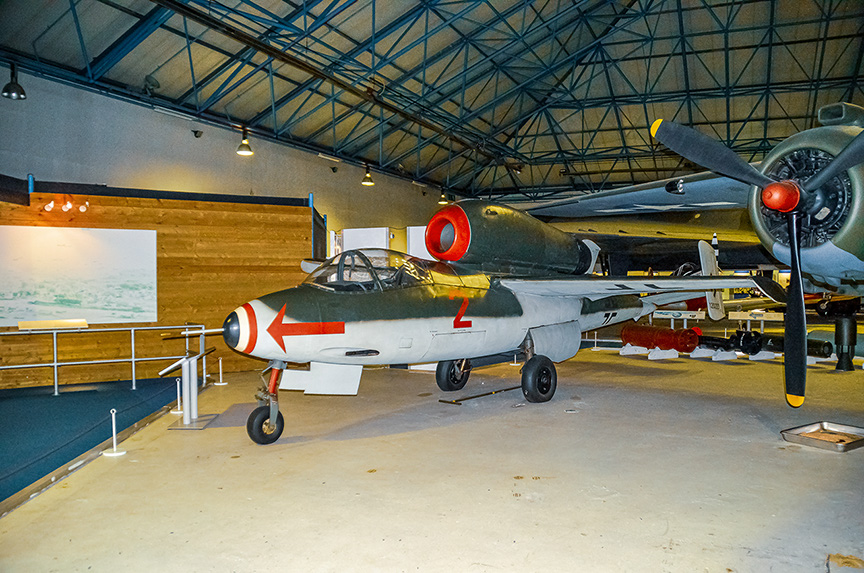 The image depicts a vintage military aircraft displayed in a museum setting. The aircraft is painted in a camouflage pattern with distinctive red and white markings, including a large red circle with a white cross on the fuselage and a red circle on the nose. The aircraft is positioned on a concrete floor with a small barrier around it, and there is a large propeller at the front. In the background, there is another aircraft with a similar design but different markings. The setting appears to be an indoor hangar with a high ceiling and wooden walls.