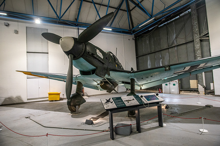 The image shows a vintage military aircraft displayed in a museum or hangar setting. The aircraft is mounted on a stand, with its propeller and other details clearly visible. There are informational displays in front of the aircraft, providing context and information about the exhibit. The setting appears to be indoors, with a high ceiling and industrial lighting. The overall atmosphere is one of historical preservation and education.