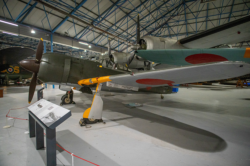 The image shows a museum exhibit featuring a vintage military aircraft with a distinctive red circle emblem on its wings, likely of Japanese origin. The aircraft is displayed indoors, with informational placards in front of it. The plane has a propeller at the front and is mounted on a stand, suggesting it is not operational. The setting appears to be a well-lit, spacious hangar with other aircraft visible in the background.