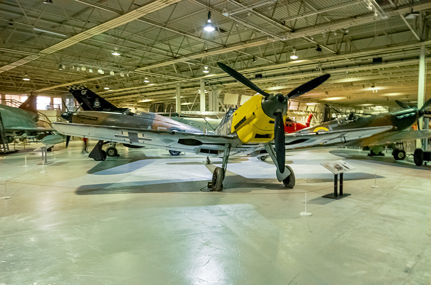 The image shows a vintage aircraft, specifically a Messerschmitt Bf 109, displayed in a museum setting. The aircraft is positioned in a large hangar with other aircraft visible in the background. The plane has a distinctive yellow nose and is mounted on a stand with an information plaque in front of it. The hangar has a high ceiling with exposed beams and industrial lighting.