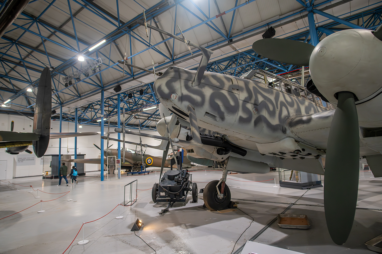 The image depicts an aircraft hangar with several vintage military planes on display. The hangar has a high ceiling with blue support beams and is well-lit. The aircraft in the foreground appears to be a World War II-era fighter plane with distinctive camouflage paint. There are a few people walking around, and the area is cordoned off with red ropes to guide visitors along a specific path.