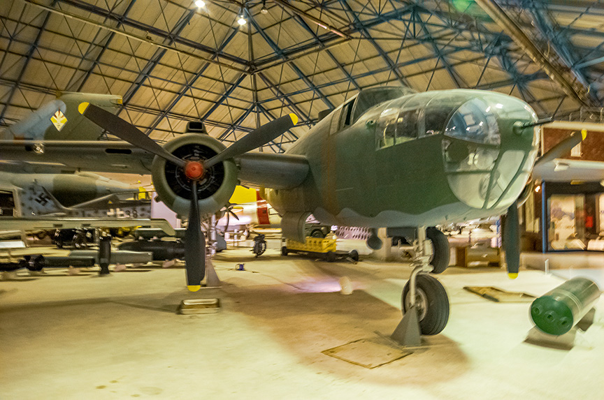 The image depicts a vintage military aircraft housed in a large hangar. The aircraft, painted in a matte green color, features a large propeller at the front and is equipped with landing gears. The hangar has a high, arched ceiling with exposed metal beams and houses other aircraft and various aviation artifacts in the background.