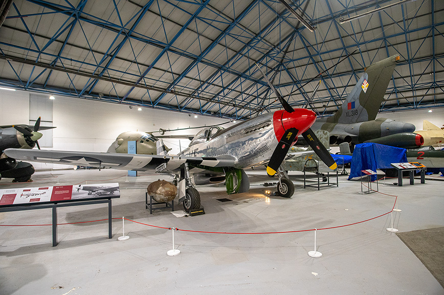 The image depicts an indoor aviation museum with several vintage military aircraft on display. The aircraft are positioned in a spacious, well-lit hangar with a high ceiling. Each plane is surrounded by informational displays and barriers to keep visitors at a safe distance. The aircraft vary in size and design, showcasing different models and eras of military aviation. The overall setting is clean and organized, providing an educational and historical overview of aviation history.