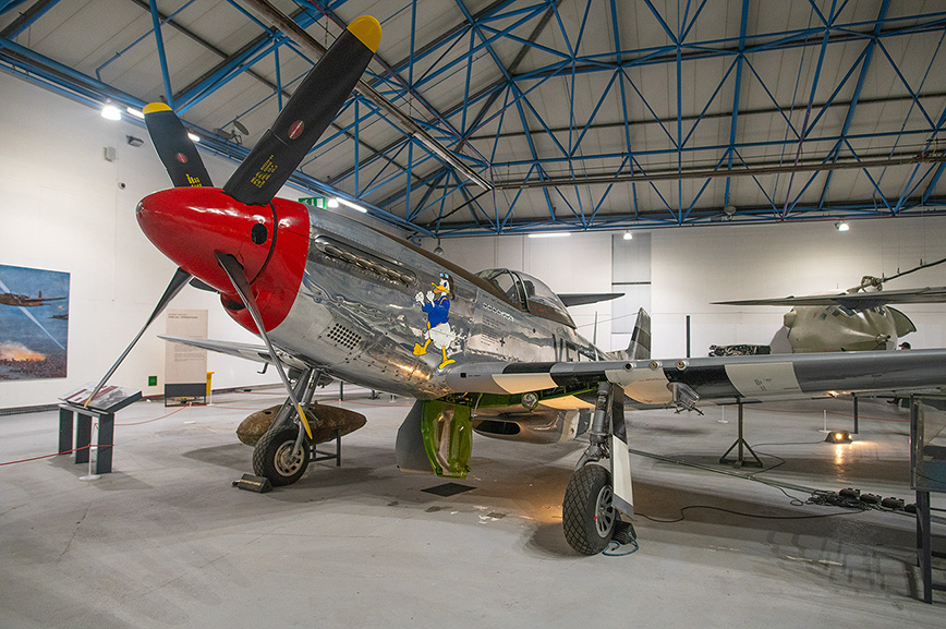 The image depicts a vintage aircraft displayed in a museum setting. The aircraft, with a distinctive red nose and silver body, is adorned with various markings and artwork. It is positioned on a concrete floor within a spacious hangar-like structure with a high, blue-trussed ceiling. Other aircraft parts and displays are visible in the background, indicating a collection of aviation artifacts.
