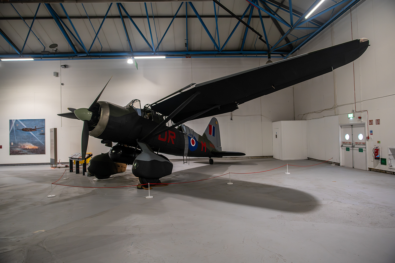 The image depicts a vintage military aircraft on display in a museum or exhibition hall. The aircraft is a single-engine, propeller-driven fighter plane, painted in a dark green color scheme with distinctive roundels and markings on its fuselage and wings. The plane is equipped with two large bombs beneath its wings. The setting appears to be an indoor hangar with a high ceiling, illuminated by overhead lights. The aircraft is cordoned off with red ropes and stanchions, indicating it is part of an exhibit.
