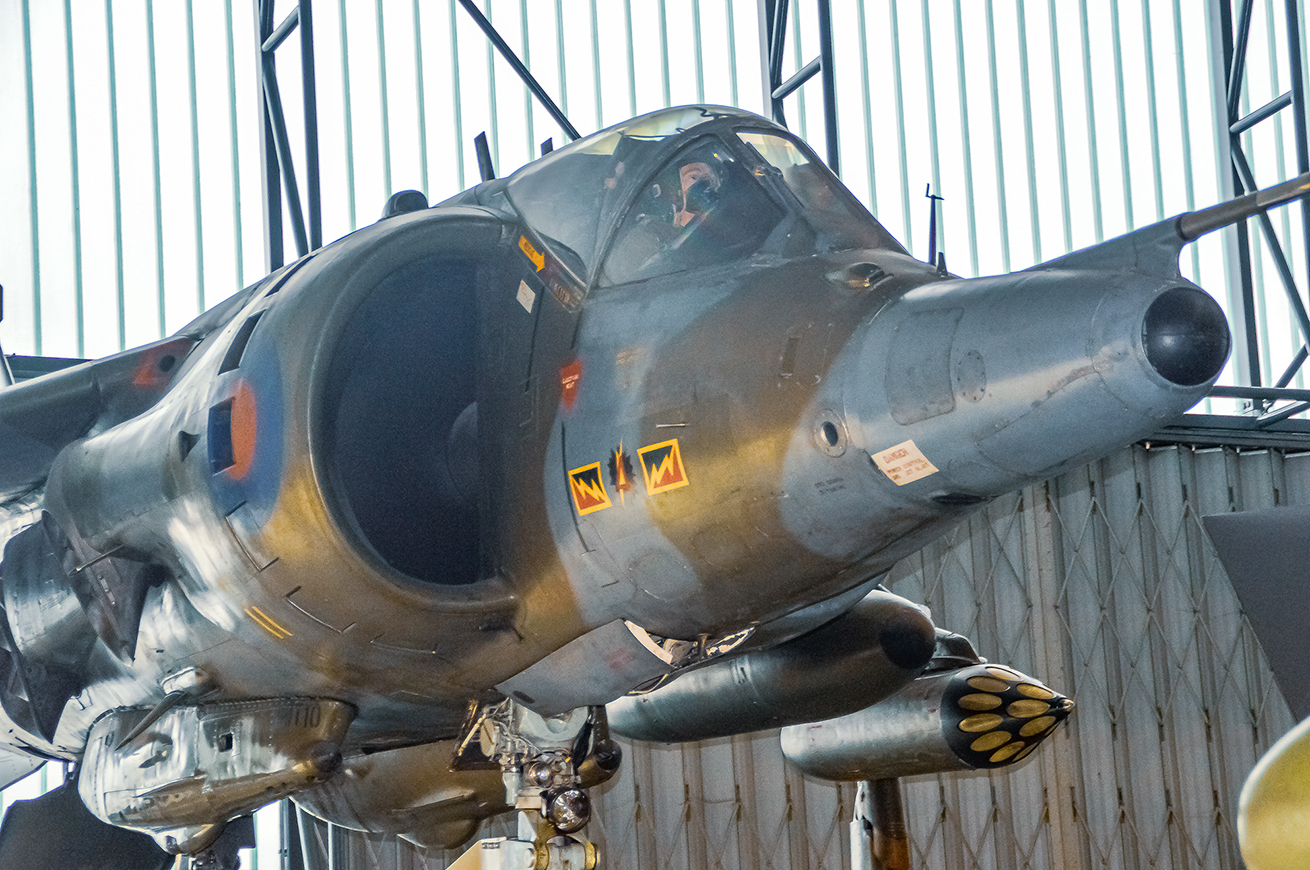 The image shows a close-up view of a military aircraft, specifically a SEPECAT Jaguar, which is a ground attack aircraft. The aircraft is displayed in a museum setting, evident from the indoor environment and the informational labels on the aircraft. The Jaguar has a distinctive nose and is equipped with various armaments and equipment. The aircraft's landing gear is down, and it is supported by a stand. The cockpit is visible, but the interior details are not clear.
