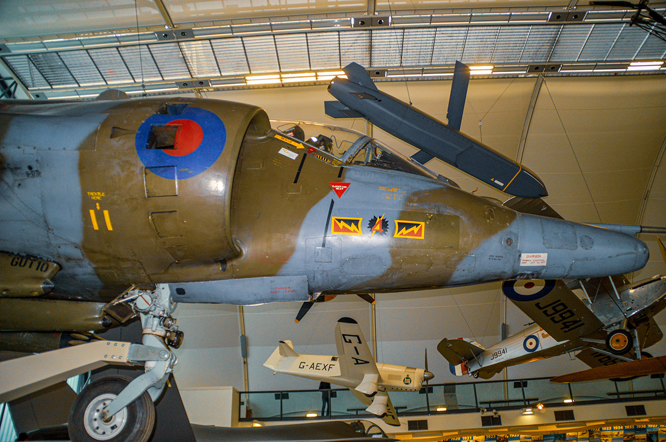 he image depicts a collection of vintage military aircraft displayed in a museum setting. The aircraft are suspended from the ceiling, showcasing their undersides and various markings. The prominent aircraft in the center features a roundel with red, white, and blue colors, along with other insignia and text. The setting appears to be a well-lit, spacious hangar with other aircraft visible in the background.