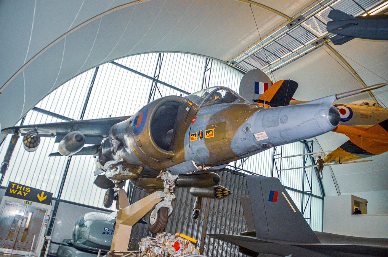 The image shows a military aircraft displayed in a museum. The aircraft is suspended from the ceiling, showcasing its underside and various markings. The setting appears to be an aviation museum with large windows and other aircraft visible in the background. The aircraft has distinct insignia and appears to be from a historical period, likely used in combat. The display is part of an exhibit, as indicated by the directional signage.