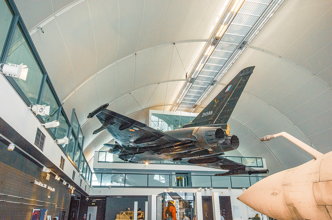 The image depicts an aircraft suspended from the ceiling in a museum-like setting. The aircraft has the tail number ZH588 and features various markings and insignia. The setting appears to be an aviation museum, with other aircraft parts and exhibits visible in the background. The environment is well-lit with natural light streaming in through large windows.