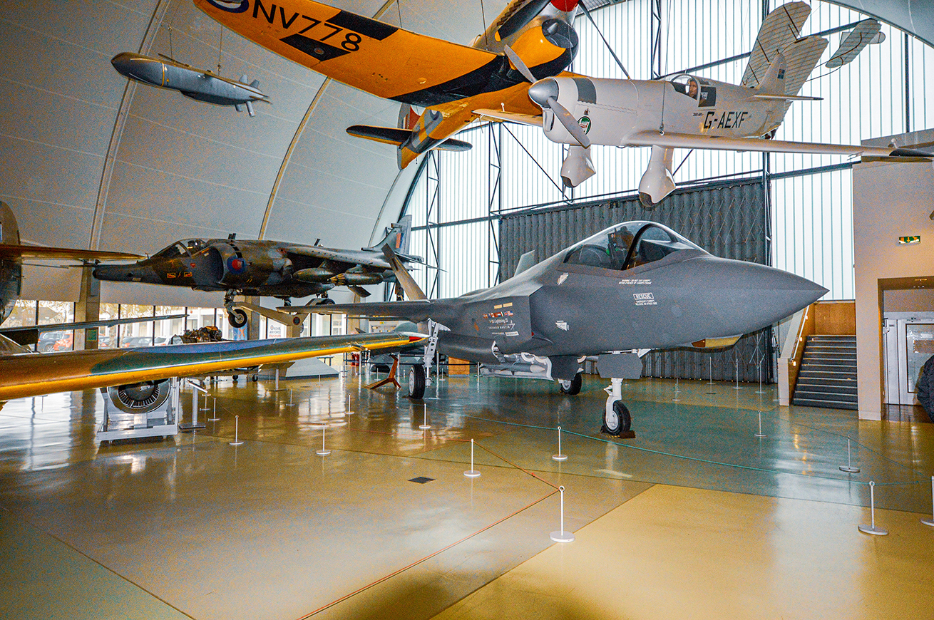 The image shows an aircraft museum with several planes on display. The planes are suspended from the ceiling and positioned on the floor, showcasing various models and designs. The setting appears to be well-lit with a polished floor, and there are small white posts on the floor, likely to guide visitors and protect the exhibits.