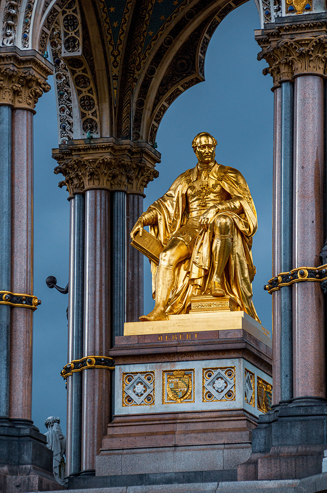The image depicts a golden statue of a seated figure, likely a notable historical person, situated under an ornate archway with intricate designs and columns. The statue holds an object in its left hand and is placed on a pedestal with detailed patterns and inscriptions. The setting appears to be part of a grand architectural structure, possibly a monument or memorial.