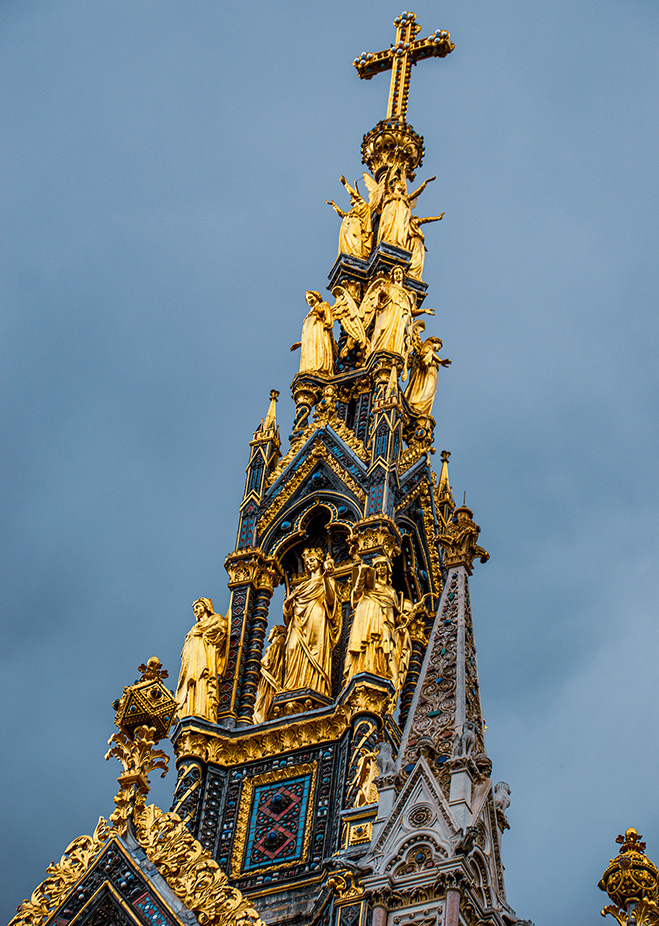 The image depicts the intricate and ornate spire of a Gothic-style cathedral. The spire is adorned with numerous golden statues and detailed architectural elements, including a prominent cross at the top. The statues appear to be of religious figures, and the spire is decorated with a mix of gold, blue, and red colors. The sky in the background is clear, providing a stark contrast to the detailed and elaborate design of the spire.