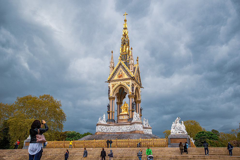 The image depicts the Albert Memorial, a grand structure located in London, UK. It is a tribute to Prince Albert, consort of Queen Victoria. The memorial features a large, ornate tower with a golden statue of Prince Albert at its center. The sky above is filled with dramatic, dark clouds, and there are people on the steps leading up to the memorial, some of whom are taking photos.
