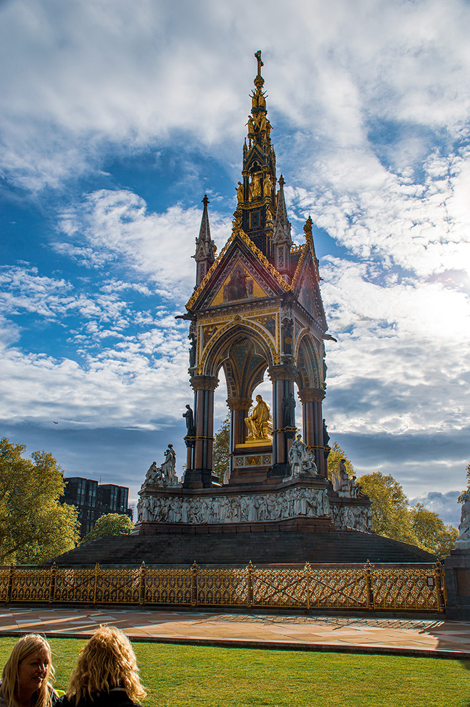 The image depicts a grand monument with intricate designs and statues, set against a partly cloudy sky. The structure features a tall spire with golden accents and multiple statues, including a prominent seated figure. The base of the monument is adorned with detailed carvings and surrounded by an ornate fence. In the foreground, two people are seated on the grass, observing the monument. The scene is set in a park-like environment with trees and modern buildings visible in the background.