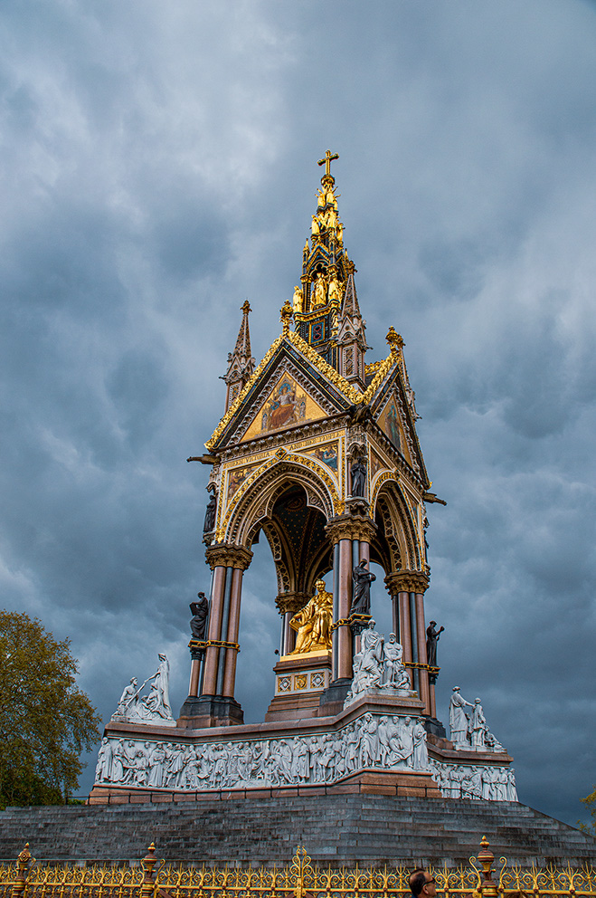 The image depicts a grand, ornate monument with intricate gold detailing and statues, set against a dramatic, cloudy sky. The structure features a central spire topped with a cross, surrounded by multiple statues and decorative elements. The base of the monument is adorned with detailed carvings and statues, and it is enclosed by a golden fence.
