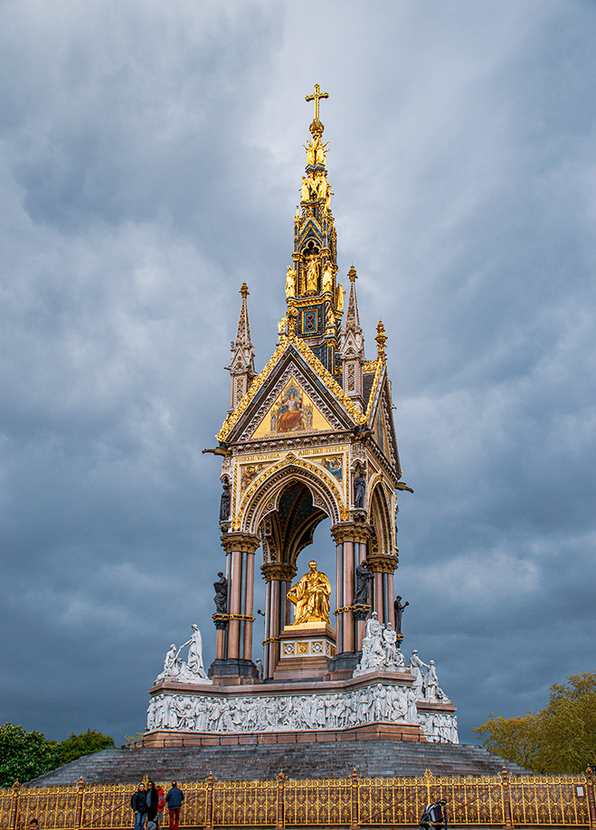 The image depicts a grand, ornate monument with a prominent golden statue at its center, set against a backdrop of dark, cloudy skies. The monument features intricate architectural details, including pointed arches, spires, and detailed carvings. The base of the monument is surrounded by a fence, and a few people are visible near the base, providing a sense of scale to the imposing structure.
