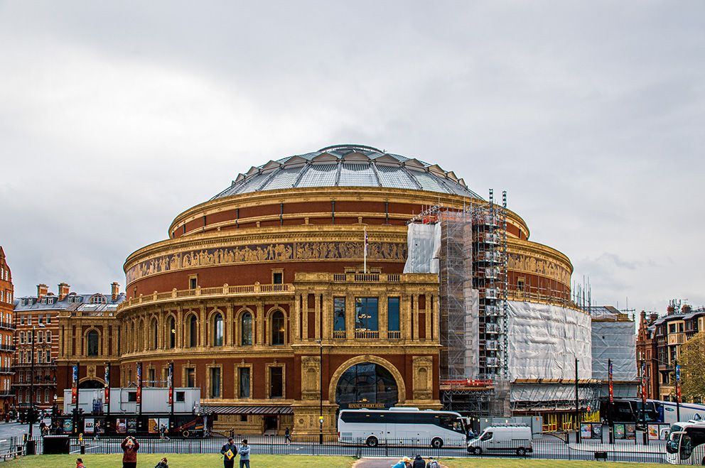 The image depicts the Royal Albert Hall, a renowned concert hall in London, undergoing renovation work. The building features a distinctive circular structure with a glass dome at the top and ornate architectural details. Scaffolding and protective coverings are visible on the right side of the building, indicating ongoing maintenance or restoration efforts. The surrounding area includes a grassy open space with a few people and vehicles present.