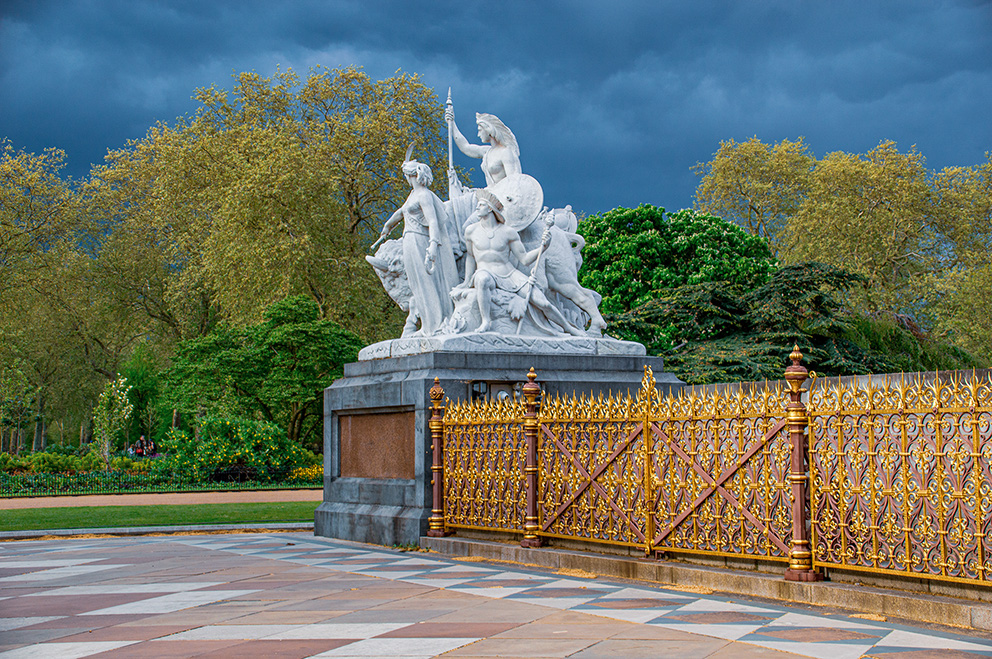 The image depicts a grand sculpture in an outdoor setting, likely a park or garden. The sculpture features multiple figures, possibly mythological or historical, with one prominent figure holding a spear and standing on a chariot pulled by horses. The sculpture is mounted on a large stone pedestal with a commemorative plaque. The area is enclosed by an ornate golden fence, and the background showcases lush greenery and a dark, cloudy sky.