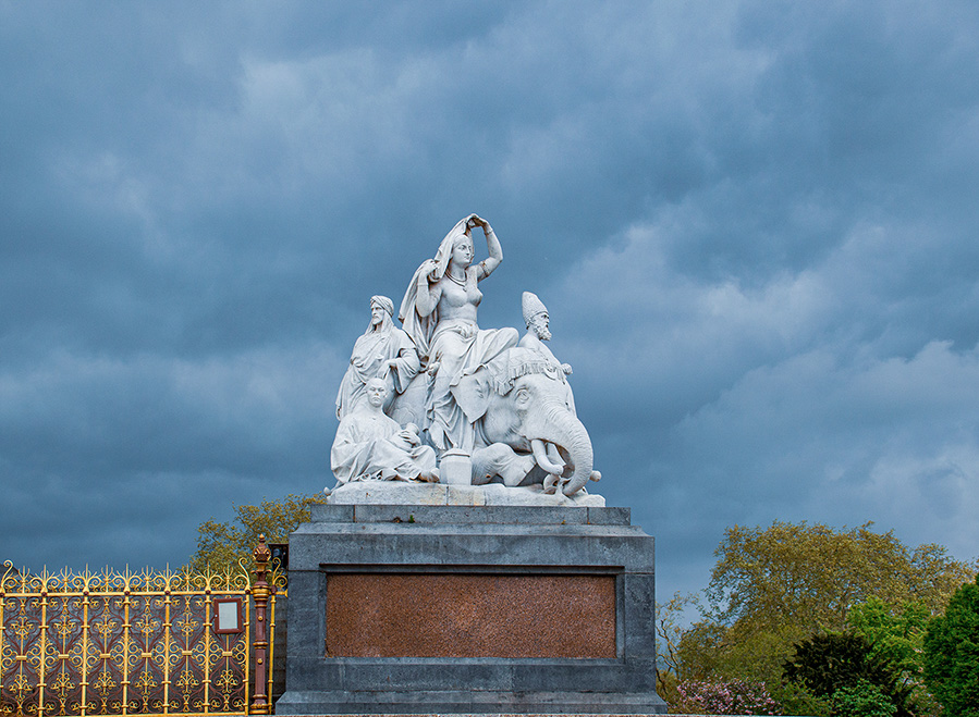 The document describes a statue featuring a central female figure with a blindfolded male figure kneeling before her, symbolizing Peace and Victory. The statue is part of a larger monument with other figures, including a seated female figure representing Justice. The monument is located in a park, surrounded by a fence, and is situated in front of a palace. The scene is set under a cloudy sky, and the statue is made of white stone, contrasting with the dark stone pedestal.