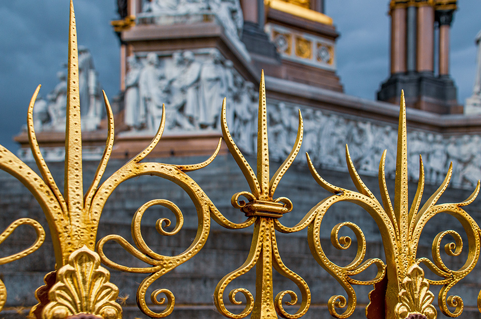 The image shows an ornate golden fence with intricate designs in the foreground, and a large, elaborate building with detailed sculptures and architectural elements in the background. The sky appears overcast, suggesting a cloudy day. The fence features pointed and curled elements, with a decorative hinge connecting two sections. The building in the background has a classical design, with columns, statues, and detailed carvings.