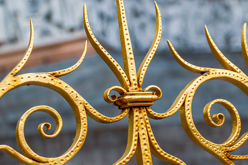 The image depicts an ornate, golden-colored metalwork design featuring intricate patterns and swirls, with a central motif resembling a stylized flower or starburst, connected by a cylindrical hinge.