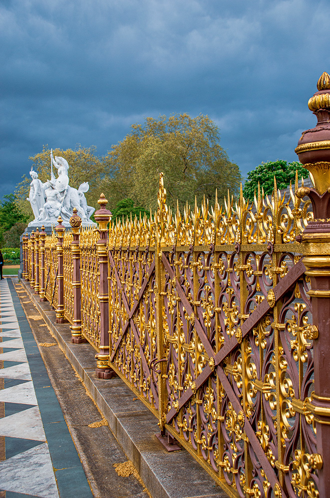 The image depicts an ornate golden fence with intricate designs and red posts topped with golden finials. Alongside the fence, there is a statue of a group of figures, possibly mythological, with one figure holding a spear. The scene is set against a backdrop of lush green trees under a cloudy sky, and the ground features a black and white checkered pattern near the fence.