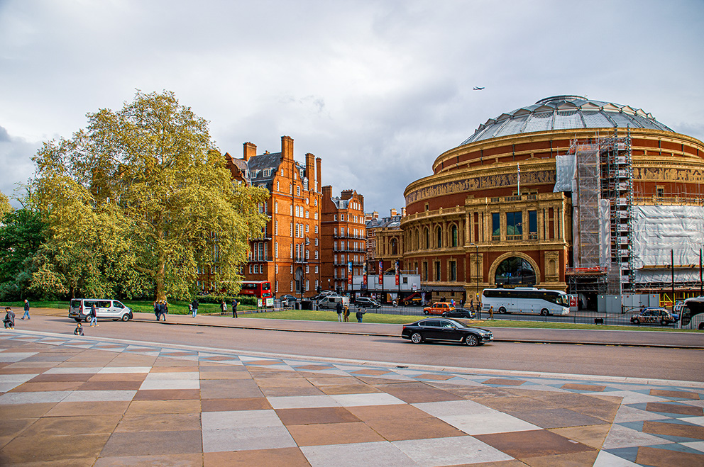 The image depicts a city scene with a prominent, ornate building under construction or renovation, likely a historical or significant structure. The building features a large dome and is surrounded by scaffolding. In the foreground, there is a spacious, patterned plaza with a few people walking and biking. Adjacent to the plaza is a park area with trees and greenery. The street adjacent to the plaza has several vehicles, including cars, buses, and a double-decker bus.
