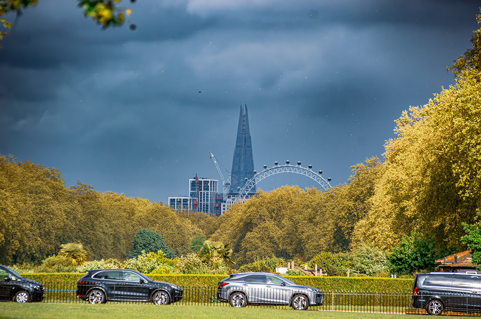 The image depicts a scenic view of a cityscape with prominent landmarks, including a tall, spire-like structure and a large Ferris wheel. The foreground features a grassy area with parked cars and a fence, while the background showcases a mix of trees with autumn foliage and a cloudy sky. The overall atmosphere suggests a blend of urban and natural elements.