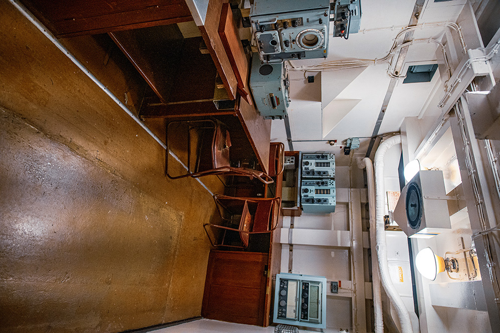 The image depicts a narrow, confined space with a steep staircase leading upwards. The walls are metallic, suggesting an industrial or maritime setting. On the left side, there are wooden benches or seats, while the right side is filled with various electronic equipment and control panels. The ceiling has exposed pipes and wiring, and there is a small, enclosed area at the bottom of the stairs that appears to be a compact bathroom.