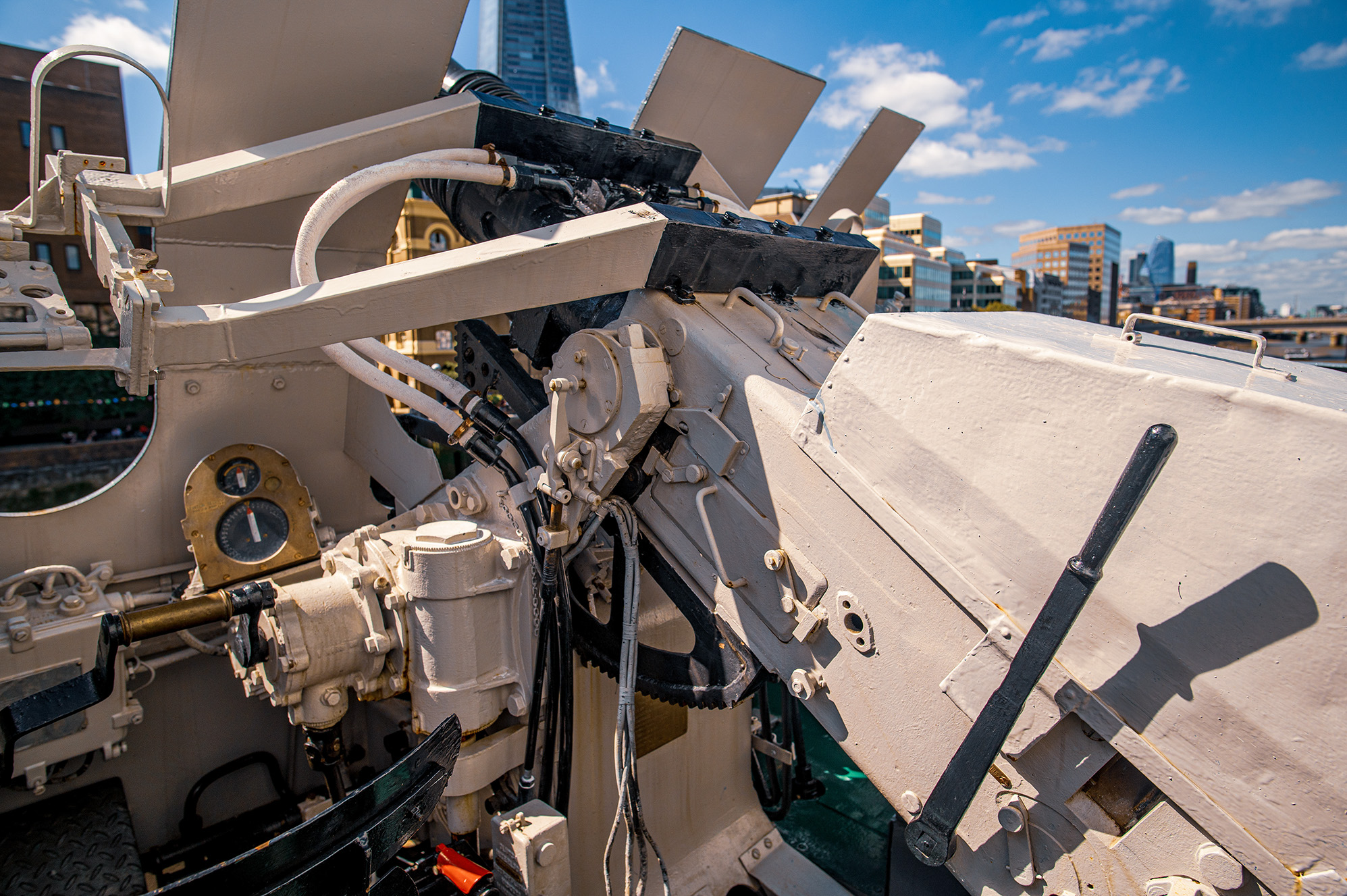 The image shows a close-up view of the turret and gun mechanism of a military tank. The tank is beige and black, with various mechanical components and instruments visible, including a pressure gauge, hydraulic systems, and control levers. The background features a cityscape with modern buildings under a partly cloudy sky.
