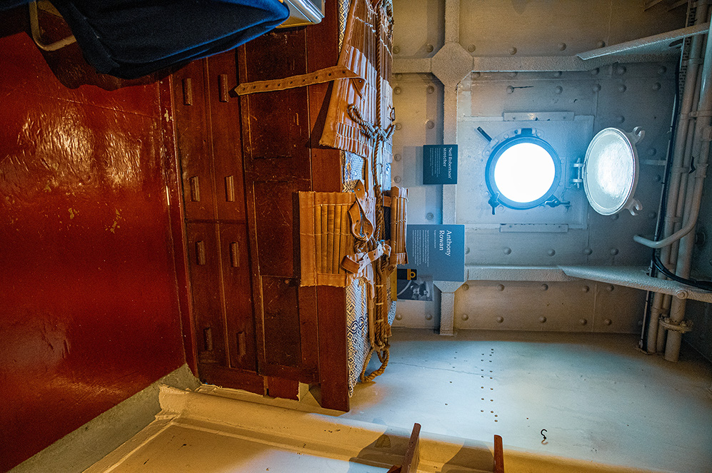 The image depicts the interior of a ship, specifically a section with a wooden door and a metal bulkhead. The wooden door is secured with metal straps and a padlock, and there is a small informational plaque on the wall. The metal bulkhead has two round portholes that allow light to enter the space. The floor is made of metal and appears to be part of a lower deck or cargo hold. The overall setting suggests an old or historical ship, possibly a cargo or storage area.