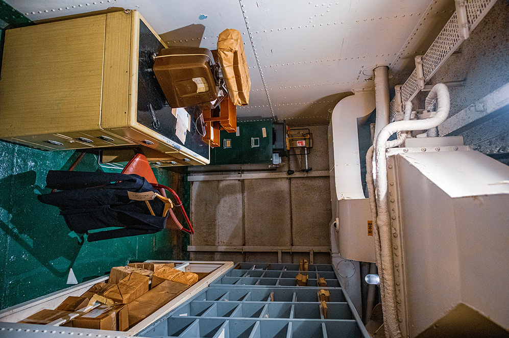 The image depicts a cluttered, narrow interior space, likely a storage or utility room. Various items, including cardboard boxes, a chair, and some personal belongings, are haphazardly placed. The room has visible pipes and a small ladder leading to a lower level. The overall appearance suggests a disorganized and possibly seldom-used area.