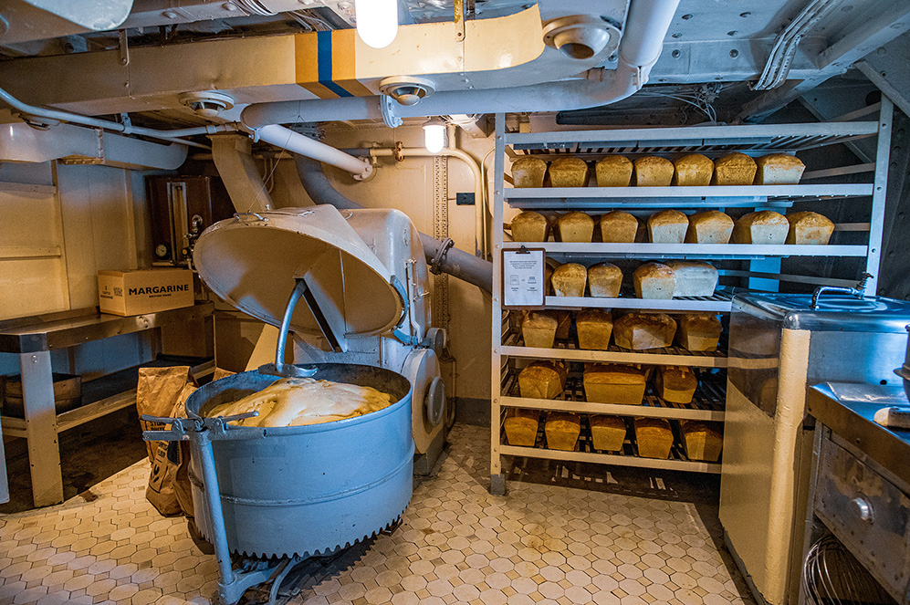 The image depicts an industrial bread-making facility with a large mixing machine and shelves filled with loaves of bread