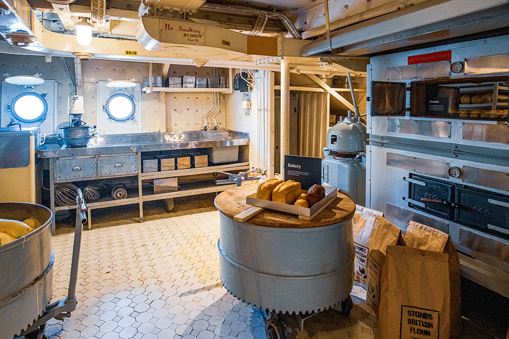 The image depicts a vintage bakery, likely on a ship, featuring old-fashioned baking equipment, including ovens, a dough mixer, and various baking utensils. The setting includes a tiled floor, metal surfaces, and porthole windows, giving it a historical and industrial feel. There are loaves of bread on display, and signs indicating 'No Smoking' and 'Bakery' are visible. The overall atmosphere suggests a well-preserved or restored historical bakery environment.